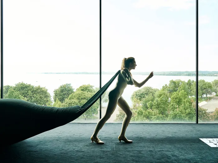 Szene aus "Ihr seid bereits eingeschifft", 2021, diverse Spielorte, Frau zieht Sack hinter sich her, vor Panoramafenster mit Blick auf Bäume und Wasser