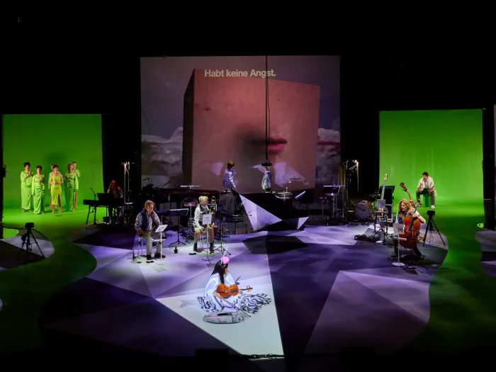 Szene aus "Melencolia", 2022 im Festspielhaus, mehrere kostümierte Darstellende und Musiker:innen auf der Bühne verteilt, große Bildschirme im Hintergrund, mittig die Aufschrift "Habt keine Angst"