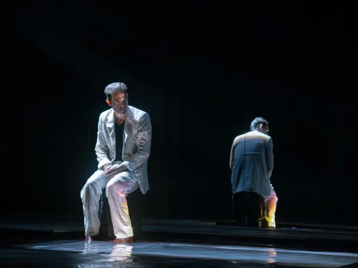 Szene aus "Der Menschenfeind", 2024 im Festspielhaus, ein sitzender Darstellender in hellem Anzug, Spiegelung der Person im Hintergrund