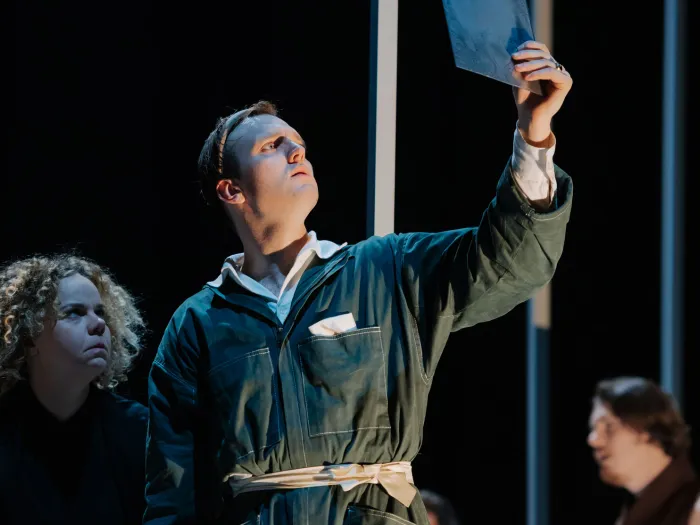 Szene aus "Richard II.", 2021 im Festspielhaus, drei Darstellende, zentral ein Darstellender in dunkelgrünem Overall mit hochgehaltenem Dokument in der Hand