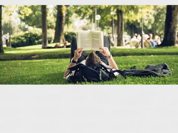 Person liegt auf dem Rücken im Park und liest ein Buch, umgeben von Bäumen und Gras