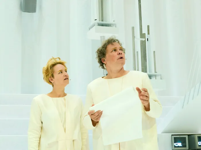 Szene aus "Wind", 2021 im Festspielhaus, zwei Darstellende in weißem Kostüm, Blick nach links richtend, eine Person hält ein großes Blatt Papier, heller Hintergrund mit Treppen