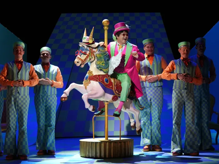 Ein Mann in einem bunten Anzug mit einem Zylinder reitet auf einem weißen Karussellpferd. Um ihn herum stehen mehrere Männer in gestreiften Westen und Krawatten.