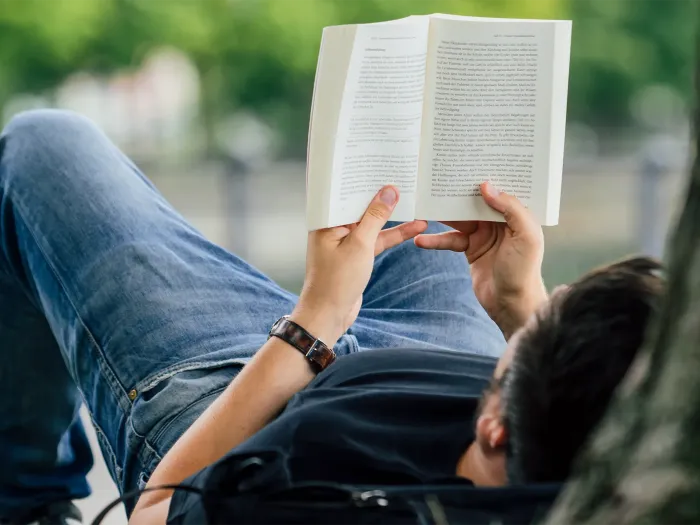 Person liegt entspannt auf dem Rücken und liest ein aufgeschlagenes Buch im Freien