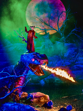 Bühnenbild von 'Der Freischütz' mit einer Figur in rotem Kostüm und Hörnern auf einem feuerspeienden Drachen vor einem großen, bunten Vollmond und düsteren, kahlen Bäumen
