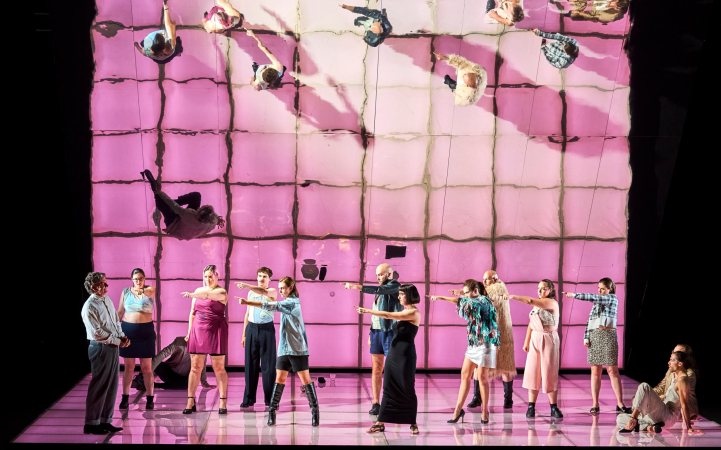 Szene aus "Die Judith von Shimoda", 2023 im Festspielhaus, zwölf kostümierte Darstellende, elf auf eine Person zeigend, rosa-farbender Boden, spiegelnde Wand im Hintergrund