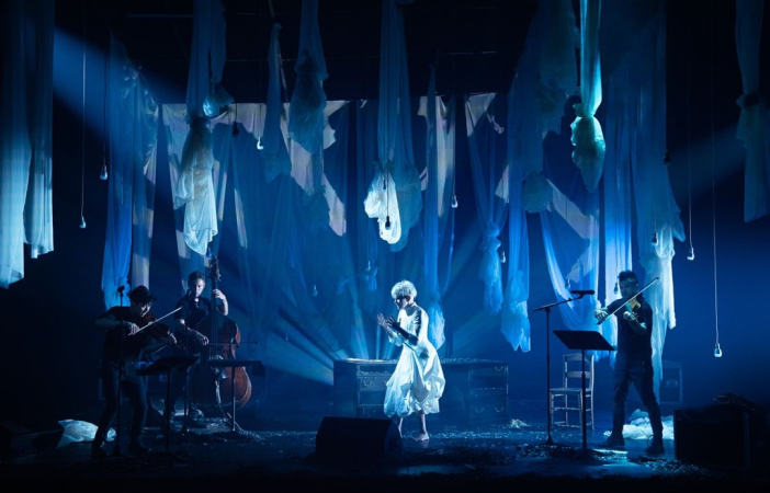 Bühnenbild mit hängenden weißen Stoffbahnen, Musiker mit Streichinstrumenten und eine Person in weißem Kostüm in der Mitte