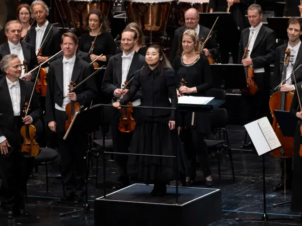 Eine Dirigentin in schwarzer Kleidung steht auf einem Podium vor einem Orchester. Die Musiker halten ihre Instrumente, darunter Geigen und Celli, und tragen formelle Kleidung.