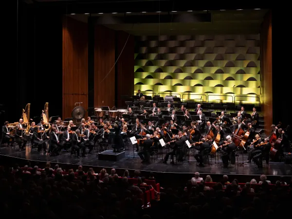 Ein Orchester mit etwa 40 Musikern in formeller Kleidung spielt auf einer Bühne. Im Hintergrund sind gestufte, grüne Wände sichtbar. Die Zuschauer sind im Vordergrund zu sehen.