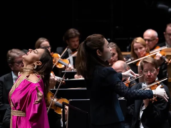 Eine Sängerin in einem pinken Kleid mit goldenen Verzierungen steht auf der Bühne und singt. Neben ihr dirigiert eine Frau in schwarzer Kleidung das Orchester im Hintergrund.