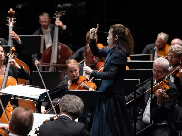 Eine Dirigentin in schwarzer Kleidung dirigiert ein Orchester. Im Hintergrund sind mehrere Musiker mit Streichinstrumenten zu sehen.