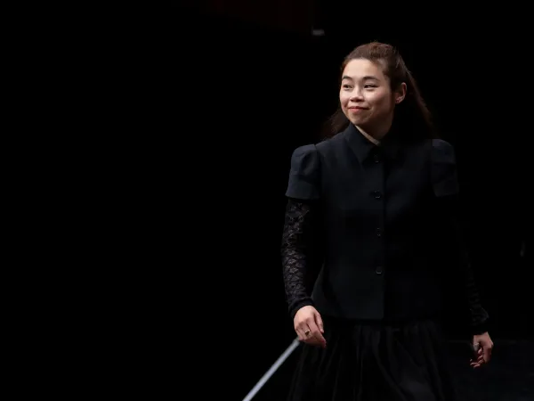 Eine Person in schwarzer Kleidung mit kurzen Ärmeln und einer langen, dunklen Rock steht auf einer Bühne. Die Person hält einen Taktstock.