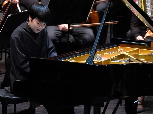 Ein Pianist sitzt an einem schwarzen Flügel mit goldenem Innenleben. Im Hintergrund sind Orchestermusiker mit Instrumenten zu sehen.