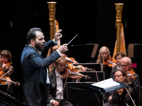 Ein Dirigent in einem schwarzen Anzug führt mit einem Taktstock vor einem Orchester. Im Hintergrund sind mehrere Musiker mit Streichinstrumenten zu sehen.