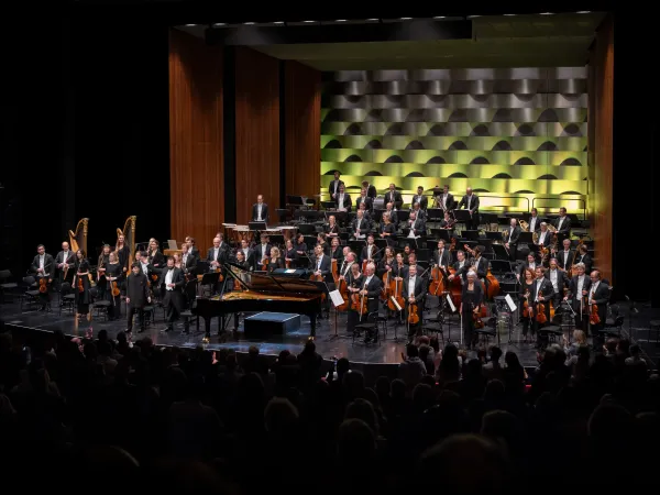 Ein Orchesterkonzert mit einem großen Orchester auf der Bühne. Im Vordergrund stehen zwei Harfen und ein Klavier. Musiker in formeller Kleidung spielen verschiedene Instrumente. Im Hintergrund sind Holz- und Blechbläser sowie Streicher zu sehen.