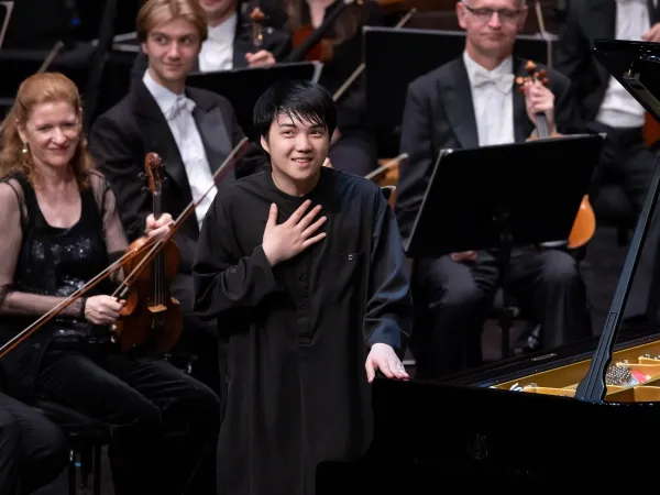 Ein Pianist in schwarzer Kleidung steht am Klavier und hält eine Hand auf der Brust. Im Hintergrund sind Orchestermusiker mit Streichinstrumenten zu sehen.