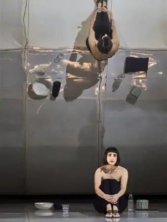 Eine sitzende Person mit langen, dunklen Haaren trägt ein schwarzes Oberteil und sitzt auf dem Boden. Über ihr ist eine reflektierte Ansicht der Person zu sehen, die auf einem glatten, reflektierenden Boden steht. Um sie herum sind verschiedene Gegenstände wie eine Schüssel, ein Glas und eine Flasche sichtbar.