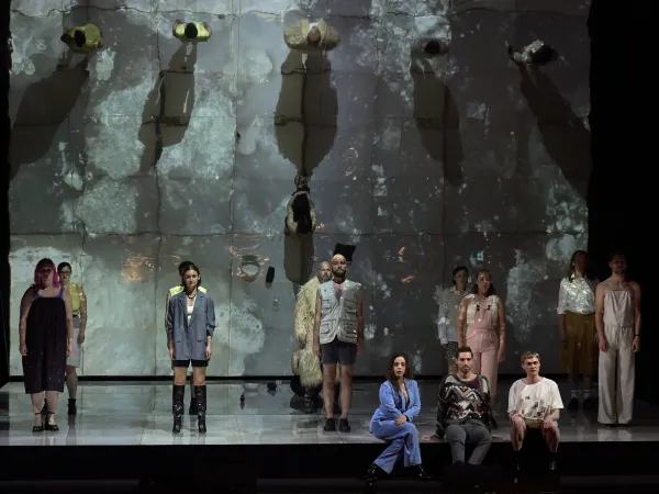Aufführung mit mehreren Personen auf einer Bühne, die in verschiedenen Outfits stehen oder sitzen. Im Hintergrund ist eine große, strukturierte Wand mit reflektierenden Oberflächen zu sehen.