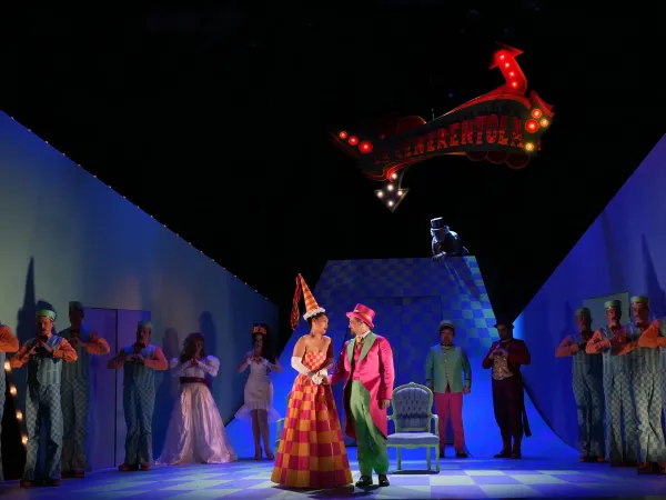 Bühnenbild einer Theateraufführung mit einer Frau in einem bunten, gestreiften Kleid und einem Mann in einem gestreiften Anzug. Im Hintergrund stehen mehrere Personen in Kostümen. Über der Szene hängt ein beleuchtetes Schild mit dem Text 'Cenerentola'.