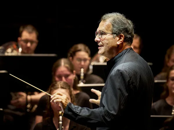 Ein Dirigent in einem schwarzen Hemd steht mit einem Taktstock in der Hand vor einem Orchester. Im Hintergrund sind Musiker mit Instrumenten zu sehen.
