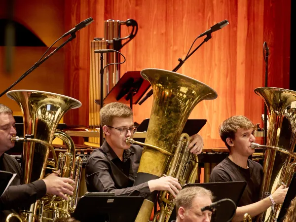 Zwei Musiker mit großen, glänzenden Blechblasinstrumenten, darunter eine Tuba, sitzen in einem Orchester. Im Hintergrund sind Mikrofone und weitere Instrumente sichtbar.