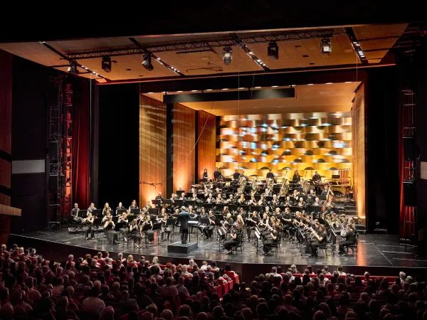 Ein Orchester mit über 50 Musikern auf der Bühne, die in einem modernen Theater spielen. Im Hintergrund ist eine farbige, geometrische Wandgestaltung sichtbar.