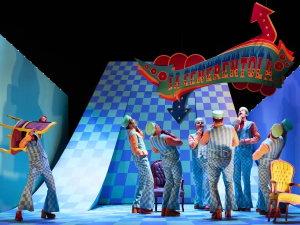 Szenenbild aus 'La Cenerentola' mit mehreren Personen in gestreiften Kostümen. Eine Person hält einen Stuhl, während andere in verschiedenen Posen stehen. Im Hintergrund ein farbenfrohes Schild mit dem Titel 'La Cenerentola'.