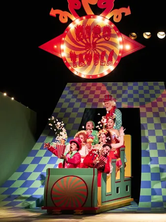 Sechs Personen in bunten Kostümen sitzen in einem grünen Wagen mit rot-grünem Muster. Im Hintergrund ein rot-weißes, spiralförmiges Schild mit 'Cenerentola'.