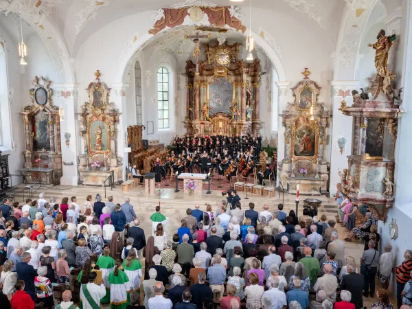 Eine große Menschenmenge steht in einer Kirche, während ein Chor und ein Orchester auf einer erhöhten Bühne auftreten. Die Kirche ist reich verziert mit Statuen und Gemälden.