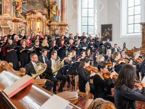 Chor und Orchester in festlicher Kleidung während einer Messe. Im Vordergrund sind Musiker mit verschiedenen Instrumenten, darunter Geigen und Hörner, zu sehen.