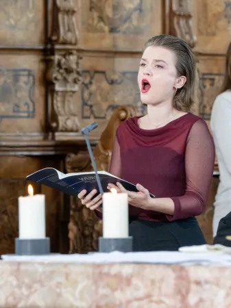 Eine Person mit schulterlangen Haaren trägt ein burgunderfarbenes Oberteil und hält ein Gesangbuch. Auf dem Tisch stehen zwei brennende Kerzen.