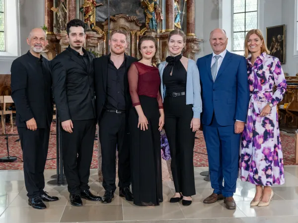 Sechs Personen stehen in formeller Kleidung in einer Kirche. Die Gruppe besteht aus drei Männern und drei Frauen, die verschiedene Farben und Stile tragen.