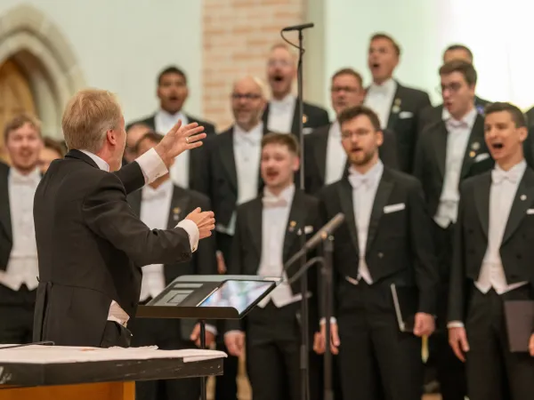 Ein Dirigent in schwarzem Anzug mit weißem Hemd und Fliege dirigiert einen Männerchor in formeller Kleidung. Der Dirigent zeigt mit der Hand auf die Sänger.
