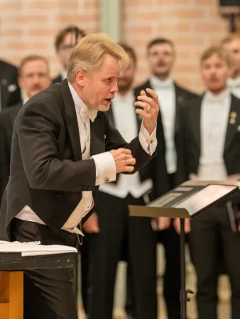 Ein Dirigent in einem schwarzen Anzug mit weißem Hemd und Fliege, der mit erhobenen Händen dirigiert. Im Hintergrund steht ein Männerchor in formeller Kleidung.