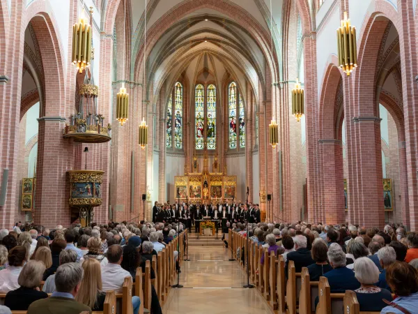 Ein gemischter Chor steht auf einer Bühne in einer Kirche mit hohen, bunten Fenstern. Das Publikum sitzt in Holzbänken und blickt nach vorne.