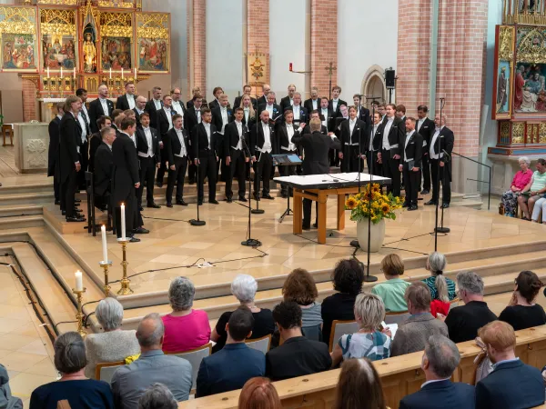 Ein gemischter Chor in formeller Kleidung singt auf einer Bühne in einer Kirche. Im Vordergrund sitzen Zuschauer auf Bänken, einige mit Notizen.