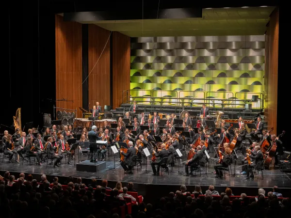Ein Orchester mit etwa 40 Musikern auf der Bühne, die Instrumente spielen. Im Vordergrund ein Dirigent, im Hintergrund eine grüne und graue Wandgestaltung.