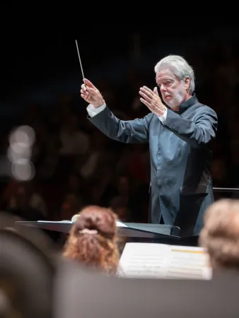 Ein Dirigent in einem schwarzen Anzug führt mit einem Taktstock. Im Hintergrund sind unscharfe Mitglieder des Orchesters und Notenblätter sichtbar.