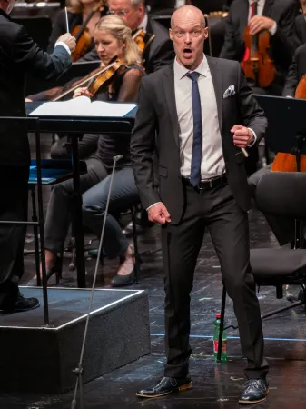 Ein Mann in einem schwarzen Anzug mit weißem Hemd und blauer Krawatte steht auf einer Bühne. Im Hintergrund sind Musiker mit Streichinstrumenten zu sehen.