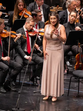 Eine Sängerin in einem schlichten, hellen Kleid steht auf der Bühne und dirigiert mit einer Hand. Im Hintergrund ist ein Orchester mit Musikern in schwarzer Kleidung und roten Krawatten zu sehen.