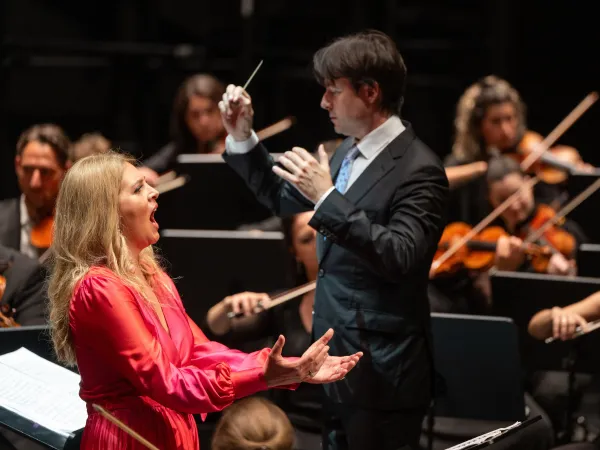 Sängerin in rotem Kleid mit ausgestreckten Armen. Im Hintergrund Dirigent mit Taktstock und schwarzem Anzug.