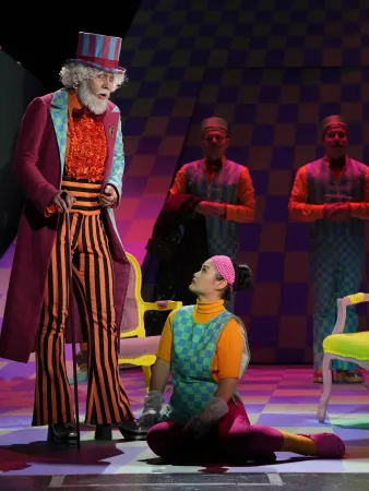 Ein Mann in einem gestreiften Anzug mit Zylinder steht vor einer knienden Person in einem bunten Outfit. Im Hintergrund sind weitere Personen in einheitlicher Kleidung sichtbar.