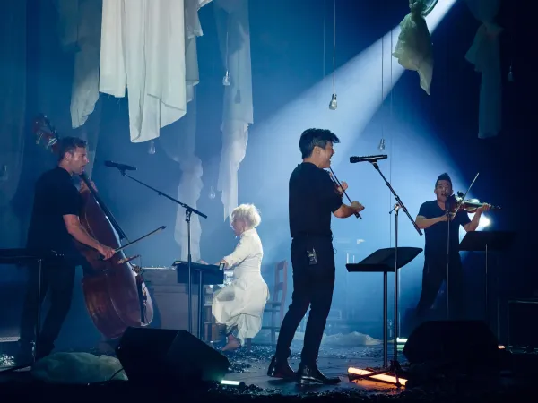 Eine Musikerin in einem weißen Kleid sitzt an einem Klavier. Um sie herum stehen drei weitere Musiker: ein Cellist, ein Violinist und ein weiterer Violinist. Die Bühne ist schwach beleuchtet und mit Stoffen dekoriert.