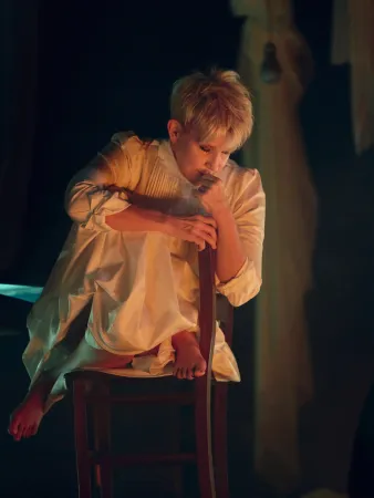 Eine Person sitzt auf einem Stuhl, trägt ein langes, helles Kleid und hat die Beine angezogen. Sie hält den Stuhl und schaut nach unten.