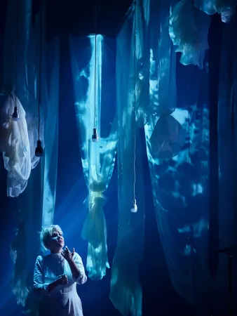 Eine Person in einem weißen Oberteil steht in einem dunklen Raum, umgeben von langen, transparenten Stoffbahnen, die von der Decke hängen. Einige Lichtquellen sind sichtbar.