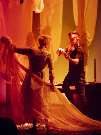 Eine Person in einem langen, transparenten Kleid hält einen Stoffvorhang, während eine andere Person im schwarzen Shirt Geige spielt. Der Hintergrund ist mit drapierten Stoffen in warmen Farben gestaltet.