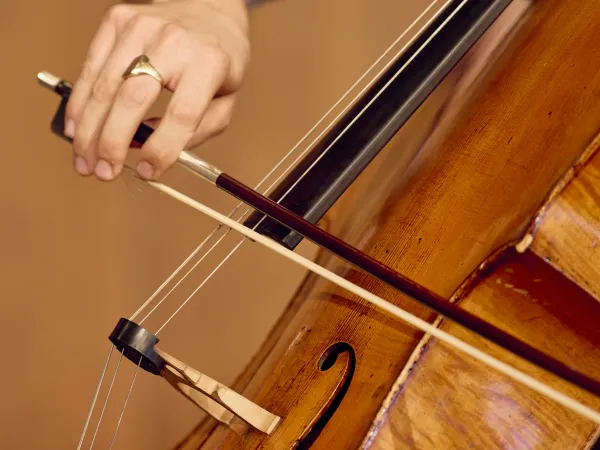 Eine Hand spielt mit einem Bogen auf den Saiten eines Cellos. Der Ring an der Hand ist sichtbar. Der Korpus des Cellos ist aus Holz.