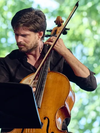 Ein Mann spielt ein Cello aus Holz. Er trägt ein schwarzes Hemd und hält den Bogen in der rechten Hand. Im Hintergrund sind grüne Bäume sichtbar.