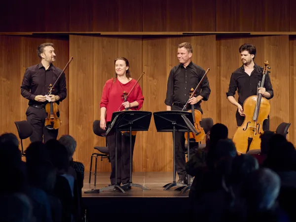Ein Streichquartett auf der Bühne. Zwei Violinisten und ein Cellist stehen in schwarzer Kleidung, eine Musikerin trägt ein rotes Oberteil. Notenständer sind vor ihnen.