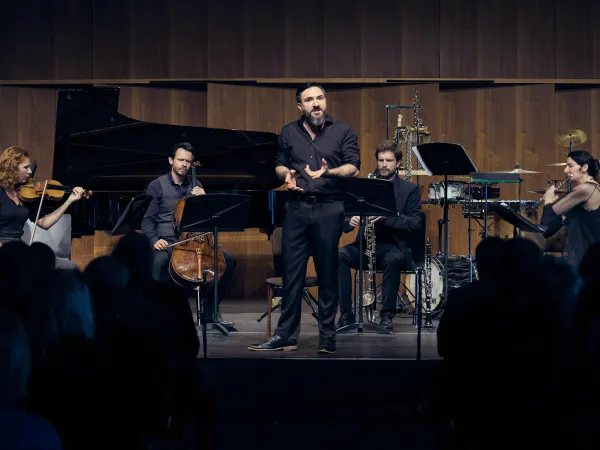 Ein Mann in schwarzer Kleidung steht im Vordergrund und dirigiert ein Ensemble. Im Hintergrund sind ein Klavier, ein Cello, eine Violine und Schlaginstrumente sichtbar. Der Saal ist mit Publikum gefüllt.